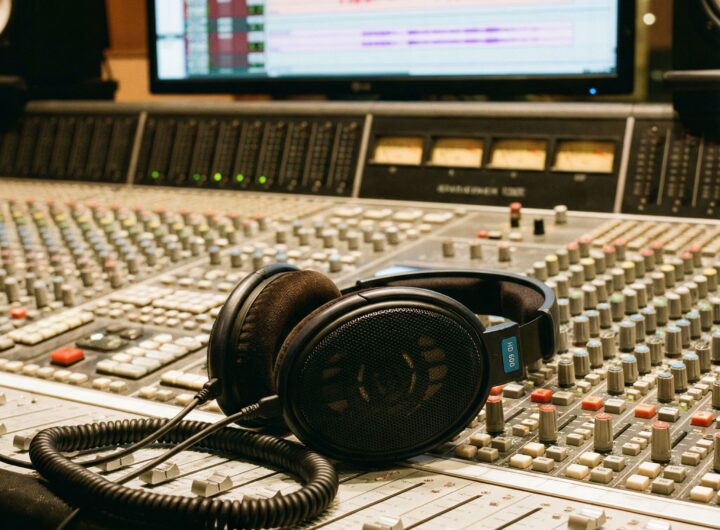 검정색 전문가용 스튜디오 헤드폰이 오디오 믹싱 콘솔 위에 놓여 있고, 배경 모니터에는 파란색 음파